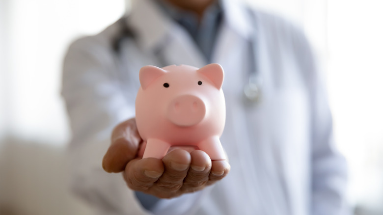 Doctor in white coat extending hand holding a small pink piggy bank