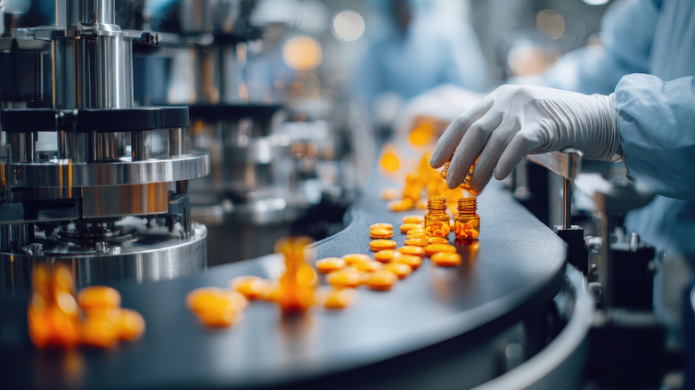 A gloved hand pouring pills into a bottle at a large manufacturing plant