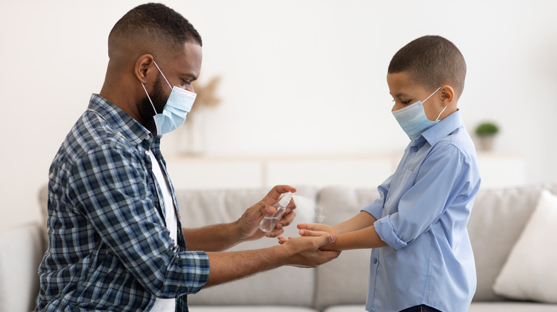 Masked father spraying hand sanitizer in hands of masked child