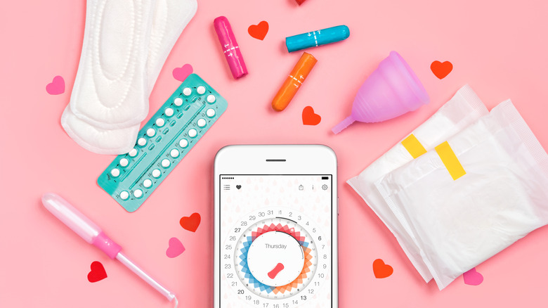 Pads, menstrual cups, and tampons surrounding a phone with a menstrual tracker open