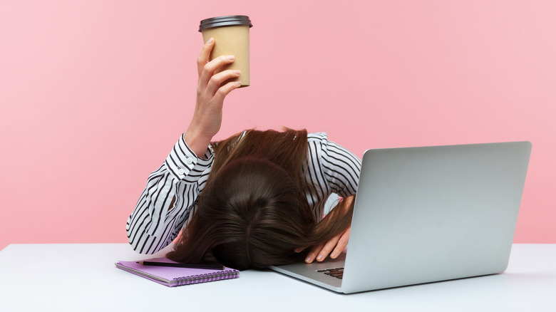 tired woman holds up coffee cup while lying head on desk