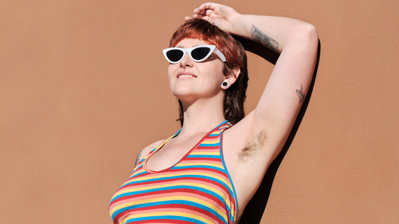 A young woman smiling and leaning against a wall while showing off her unshaven underarms in a tank top