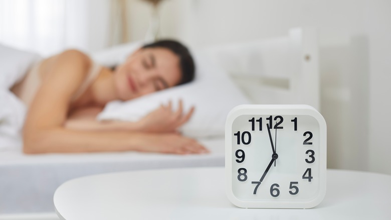 an alarm clock with a woman asleep behind it 