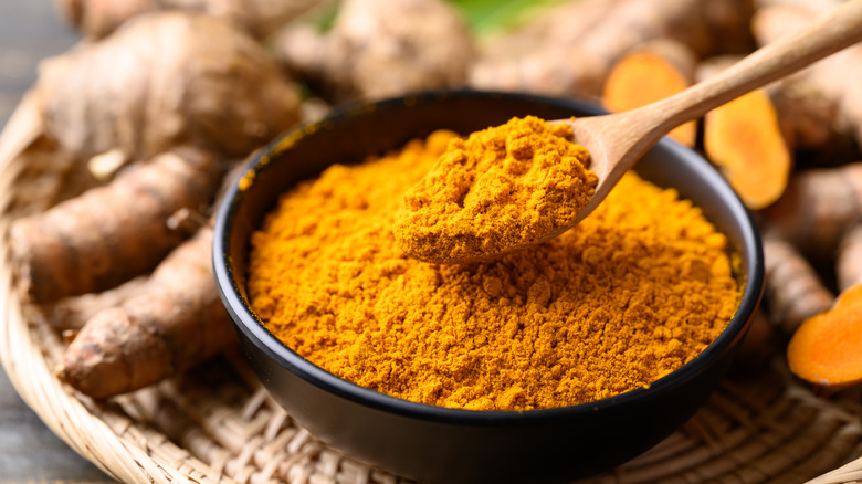 A bowl of turmeric powder with a wooden spoon surrounded by turmeric root