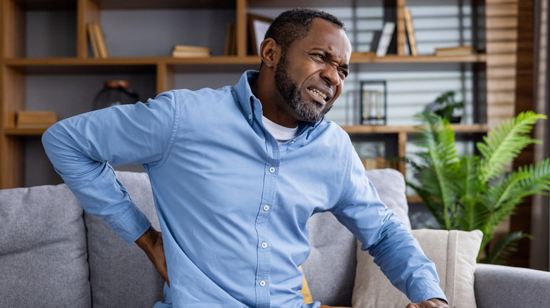 man sitting on couch holding middle of back in pain