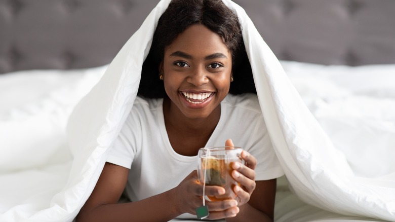 Young smiling woman popping out from under her bed covers holding a cup of tea