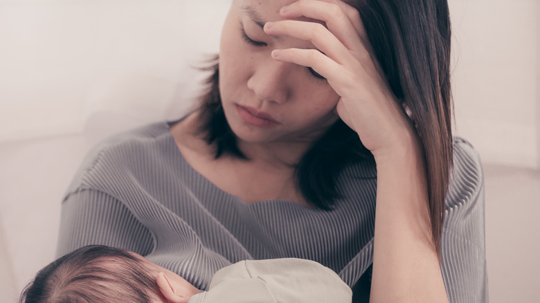 Tired mother breastfeeds young infant with head in hands