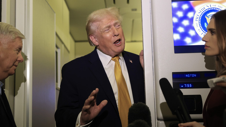 President Donald Trump with his hand raised and mouth open in conversation, surrounded by microphones and press on Air Force One.