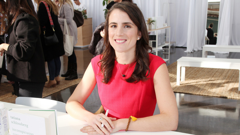 Tatiana Schlossberg sitting at a white table in a red dress, a line of people behind her as she smiles at the camera.
