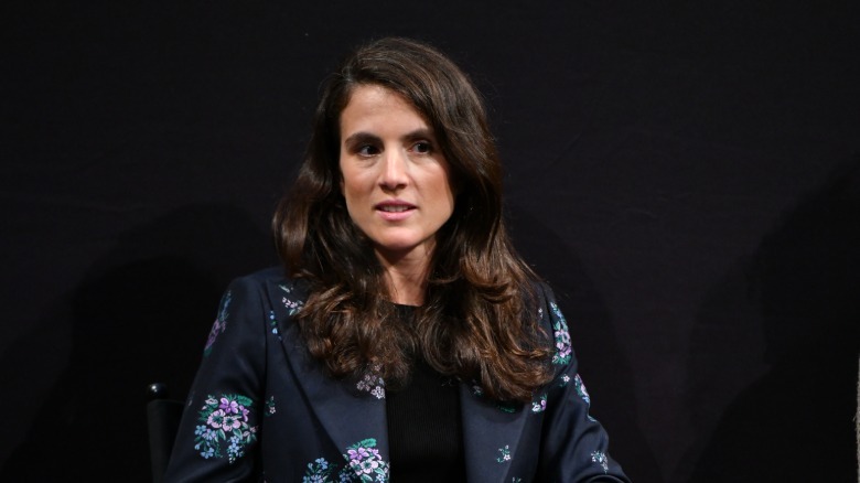 Tatiana Schlossberg sitting in a black floral outfit, holding a microphone to her mouth as she talks.