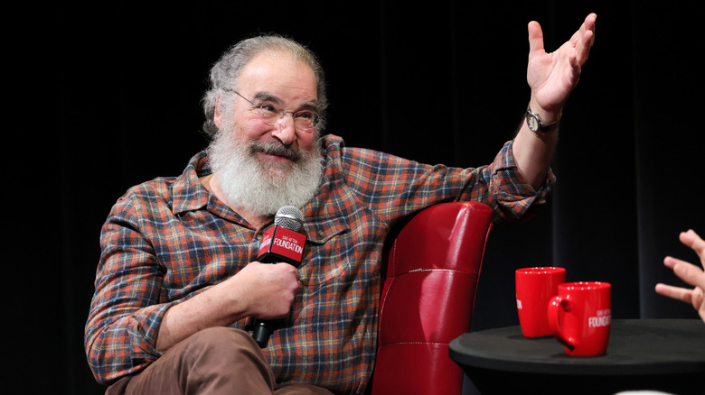 Mandy Patinkin in a plaid shirt seated in a red chair with his hand raised and a microphone in his other hand.