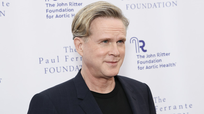 Cary Elwes in a navy blazer and black top in front of white background.