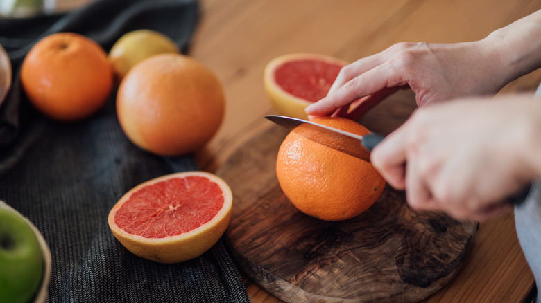 A hand slicing into an orange on a wood cutting board that's surrounded by cut grapefruit and other citrus fruit
