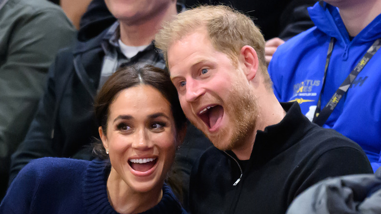 Meghan Markle and Prince Harry with their heads together making silly faces on the sideline of a basketball game.