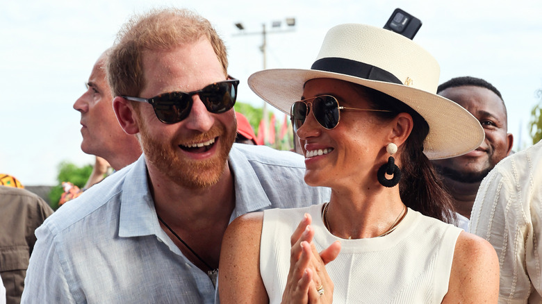 Prince Harry leaning over the shoulder of Meghan Markle, both in sunglasses.