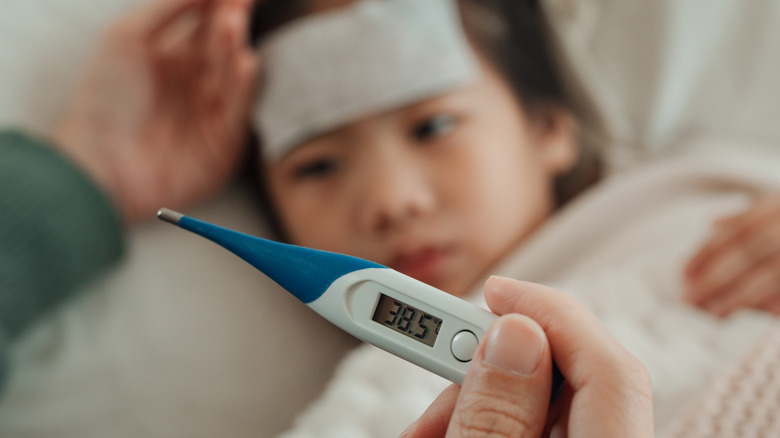 A hand holding a digital thermometer while a child with a fever lays down