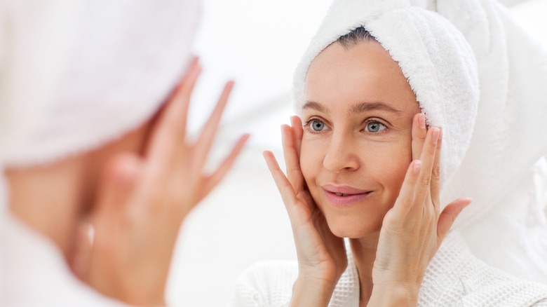 white woman in robe and towel looks at face in mirror