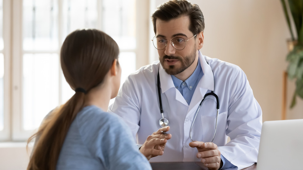 Male doctor with dark hair and glasses in lab coat with stethoscope around neck sits at desk listening to female patient who has long dark hair and is not facing camera