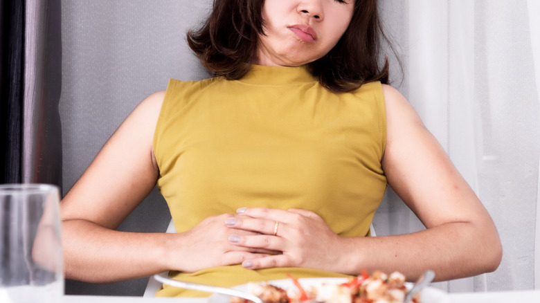 A woman leaning back from a dinner table feeling stomach bloating