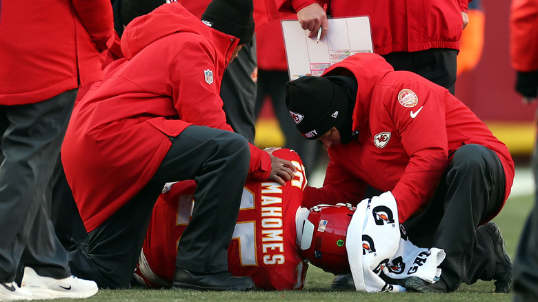 Patrick Mahomes in his football uniform laying on the ground on his side, surrounding by Chiefs staff.