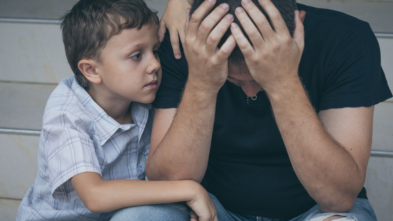 Concerned boy looks at father who sits beside him with his head in his hands