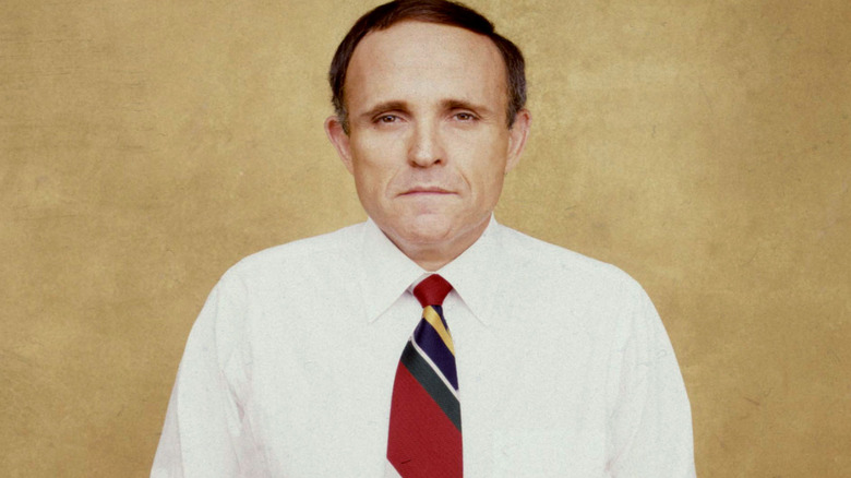 Young Rudy Giuliani in a white button up and striped tie in front of a yellow background.