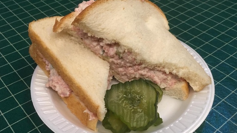 Halved bologna salad sandwich with pickle slices on white styrofoam plate