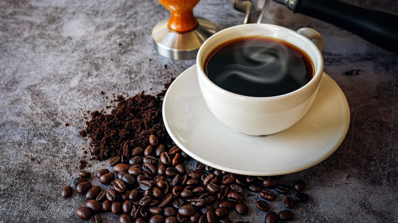 A cup of black coffee on a saucer surrounded by coffee beans and ground coffee
