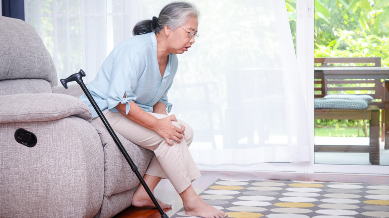 elderly woman sitting holding her knee in pain with a cane nearby