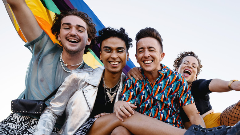 Four LGBTQ+ folks happily sit outside holding a rainbow flag overhead