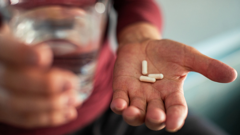 A man's hand holding three capsules and a glass of water