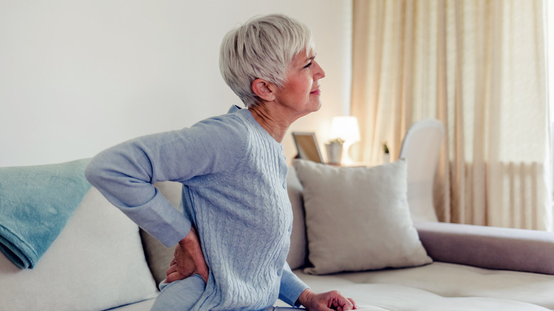 An older woman holding her lower back in pain while sitting on a couch