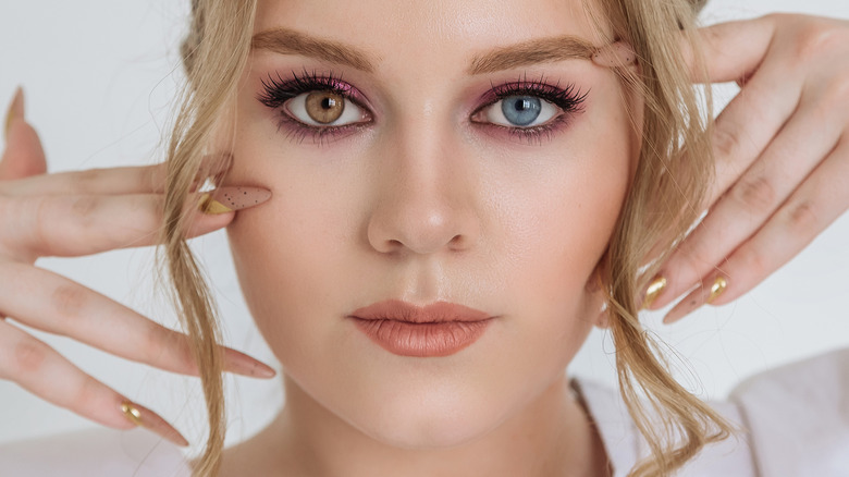 A woman with two different colored eyes posing with her manicured hands on the sides of her face
