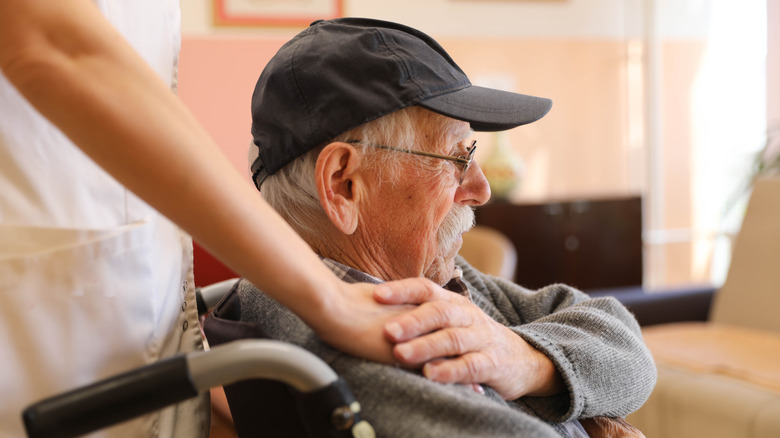 A man sitting in a wheelchair while a caregiver's hand touches his shoulder