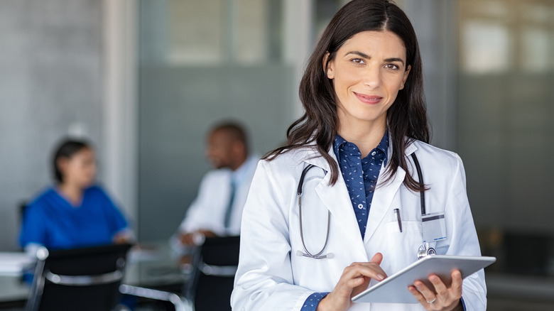 Portrait of a woman with white coat and stethoscope holding a tablet. Two people are talking in the background.