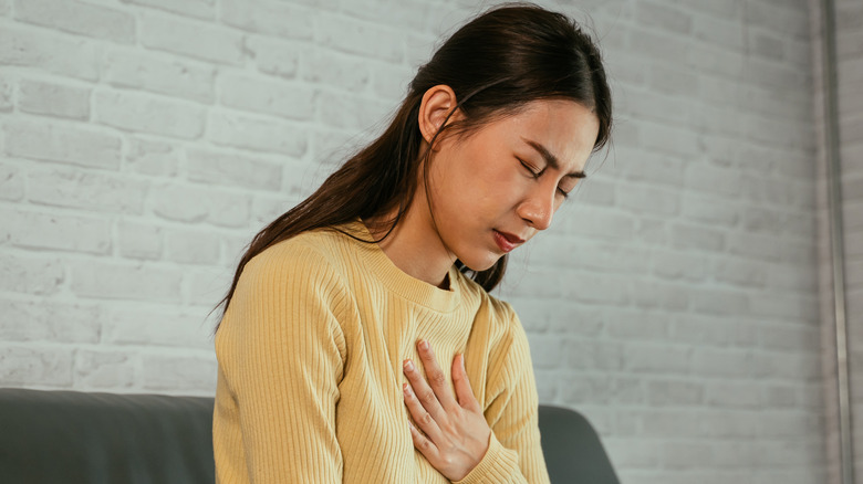 Asian young woman feeling discomfort as suffering from heartburn holding chest with closed eyes and sitting with folded legs on couch at home