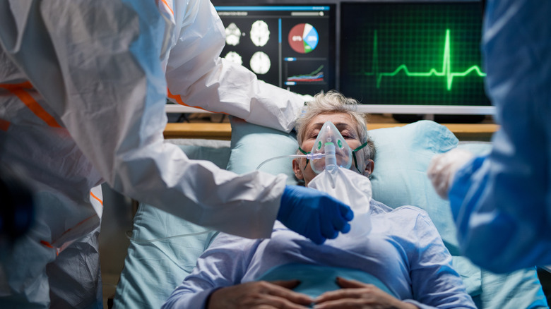 Older woman lying in hospital bed hooked up to a respirator by doctors in hazmat suits