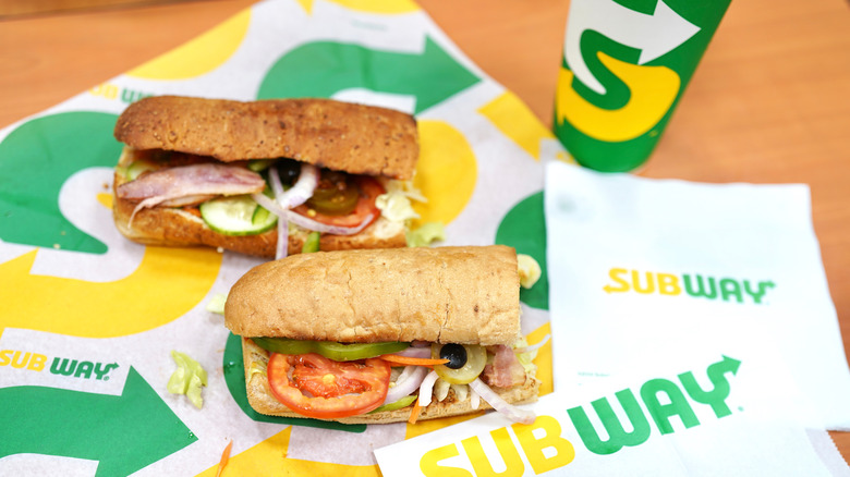 two Subway sandwich's lying on Subway paper with a fountain drink  