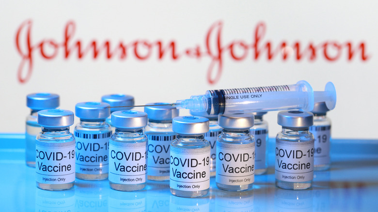 Syringe and vials of Johnson & Johnson vaccine on a table with blurred "Johnson & Johnson" logo on back wall