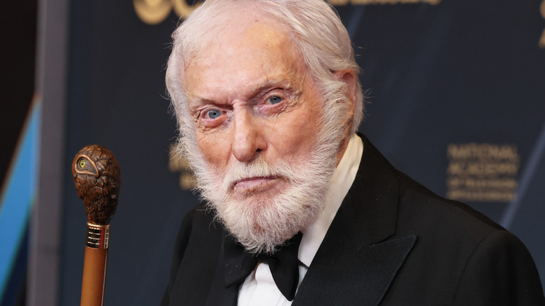 Dick Van Dyke in a tux, holding a brown, wooden cane, in front of a navy blue backdrop.
