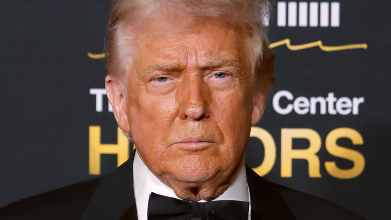 President Donald Trump, wearing a bowtie, looking directly at a camera with a black background.