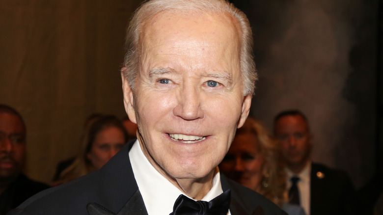 Joe Biden in a bowtie, smiling off to the left side, people fill in the space behind him.