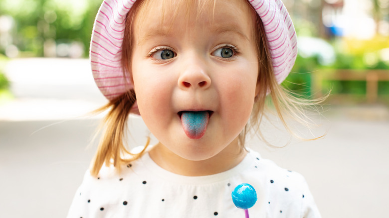 A young girl sticking out her blue tongue while holding a blue lollipop