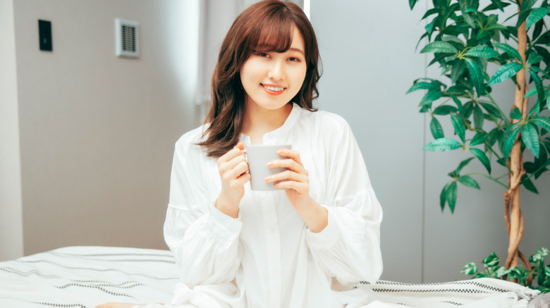 A woman enjoying a drink from a mug while sitting on her bed