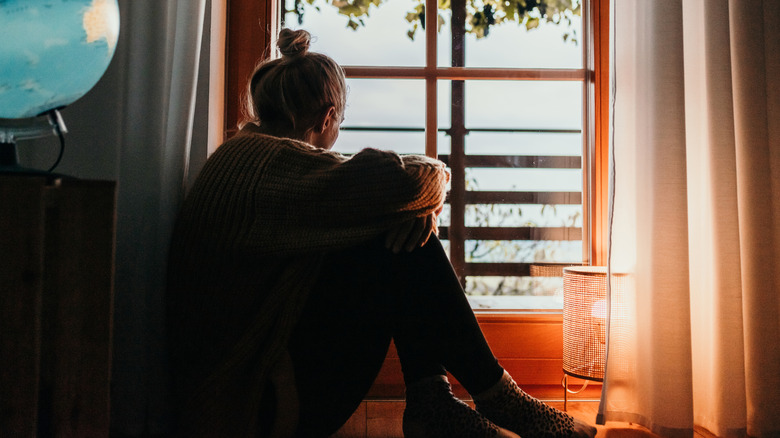 A woman sitting in her room, arms across legs, looking outside her window.