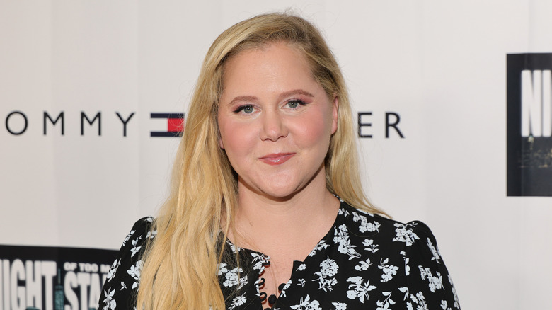 Amy Schumer in front of a white backdrop in a black floral top.
