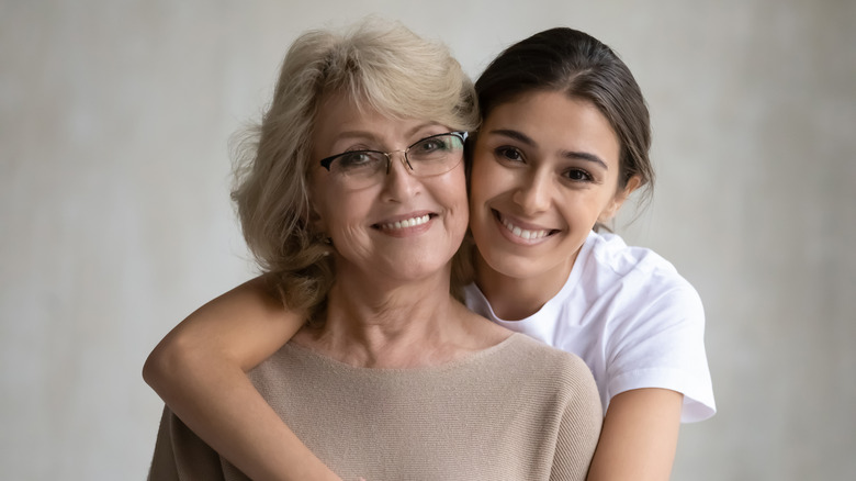 Daughter hugging middle aged mother from back, looking at camera, smiling