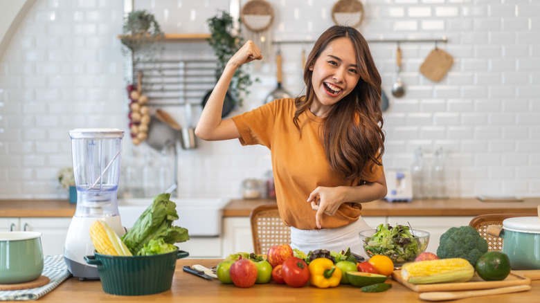 A smiling woman in her kitchen pointing to her fruits and vegetables