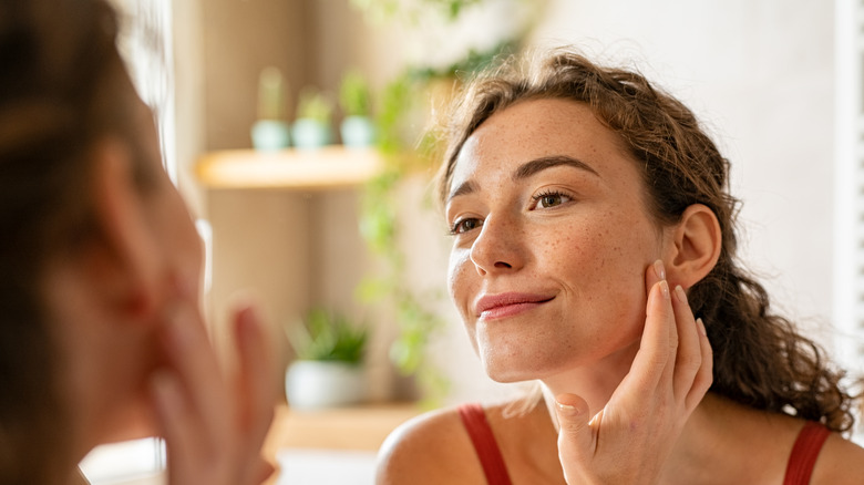 Woman looking at mirror while touching her face and checking pimple, wrinkles and bags under the eyes, during morning beauty routine.