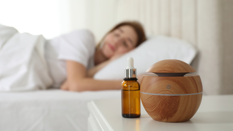 A bottle of essential oil and diffuser on a nightstand while a woman sleeps